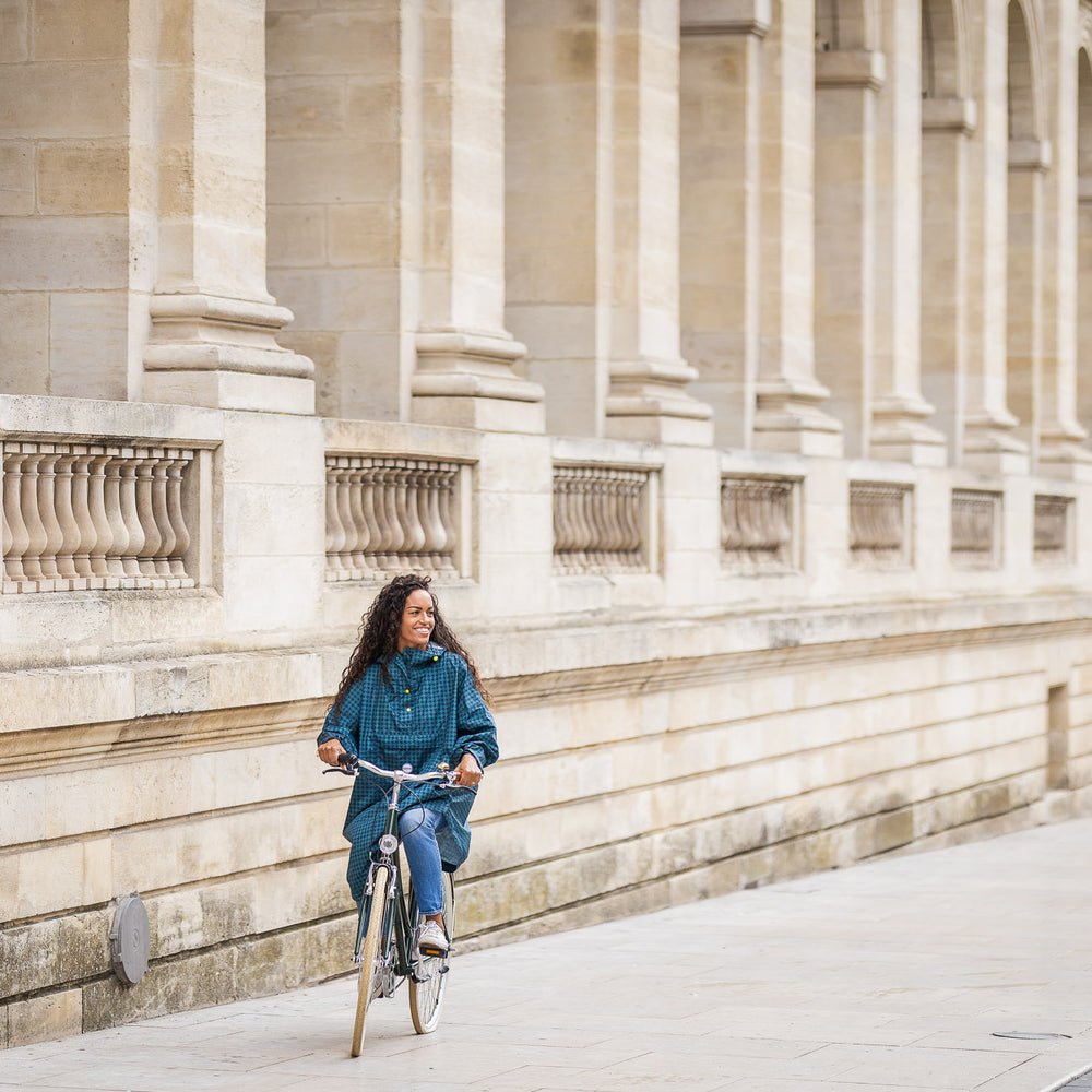 Utilisateur à vélo portant le poncho Anvers en pied de poule vert et bleu marine, coupe ample avec capuche, protection imperméable contre la pluie.