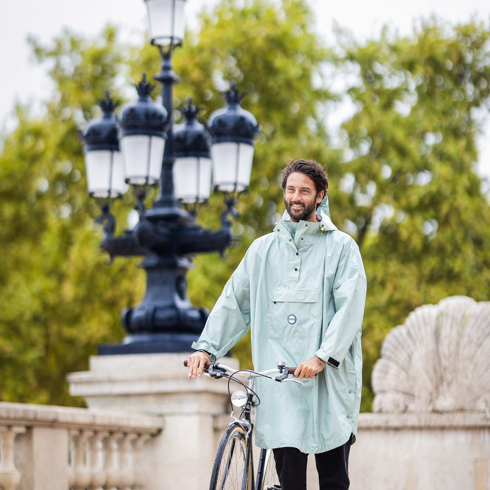 Personne à vélo portant le poncho Genève bleu glacier, sous la pluie, coupe ample avec capuche, protection imperméable.