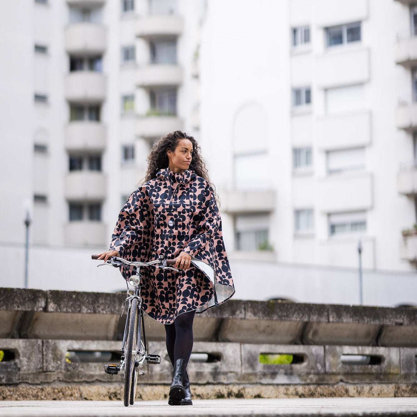 Utilisateur à vélo sous la pluie avec la cape Cap Fréhel imprimés géométriques Bronze et Noir, protection imperméable et visibilité renforcée.