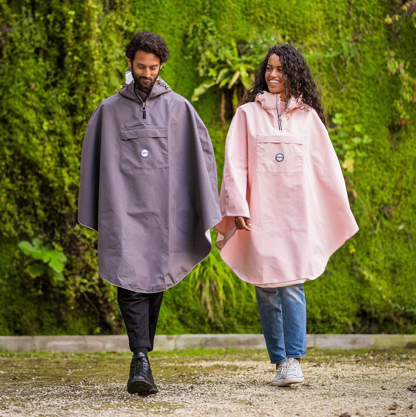 un couple portant chacun une cape de pluie rose pale et grise se promène dans un parc entouré d'un mur végétal. 