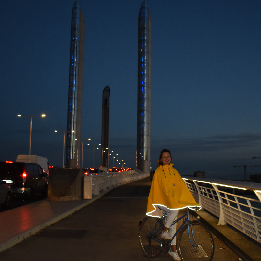 Utilisateur à vélo la nuit portant la cape Cap Breton recyclée, avec bandes réfléchissantes visibles pour une sécurité renforcée.