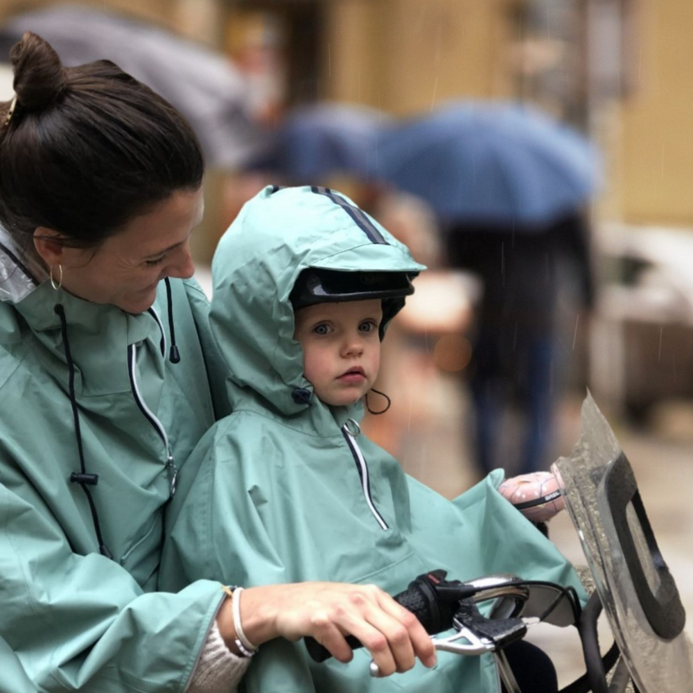 Enfant à vélo sous la pluie portant la cape de pluie 5-7 ans, protection imperméable et visibilité grâce aux bandes réfléchissantes.
